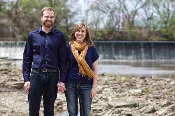 David and Lisa hold hands and stand close in the rocky shore of a river near a bridge in Bartlesville, Oklahoma